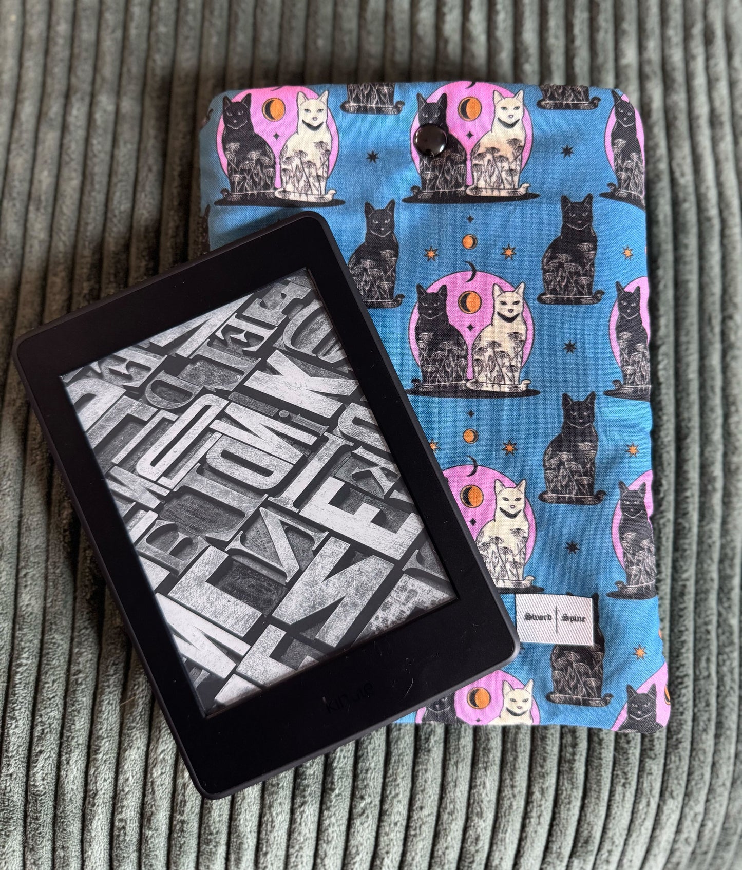 A kindle rests on a handmade padded sleeve with a cat and moon pattern in blue and pink.