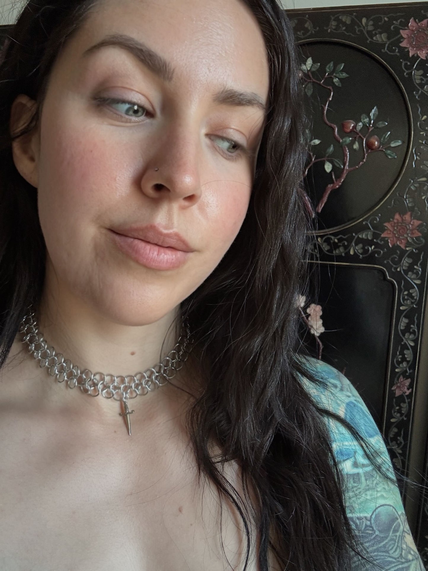 Woman wearing a silver chainmail necklace with a dagger pendant, against a decorative background.