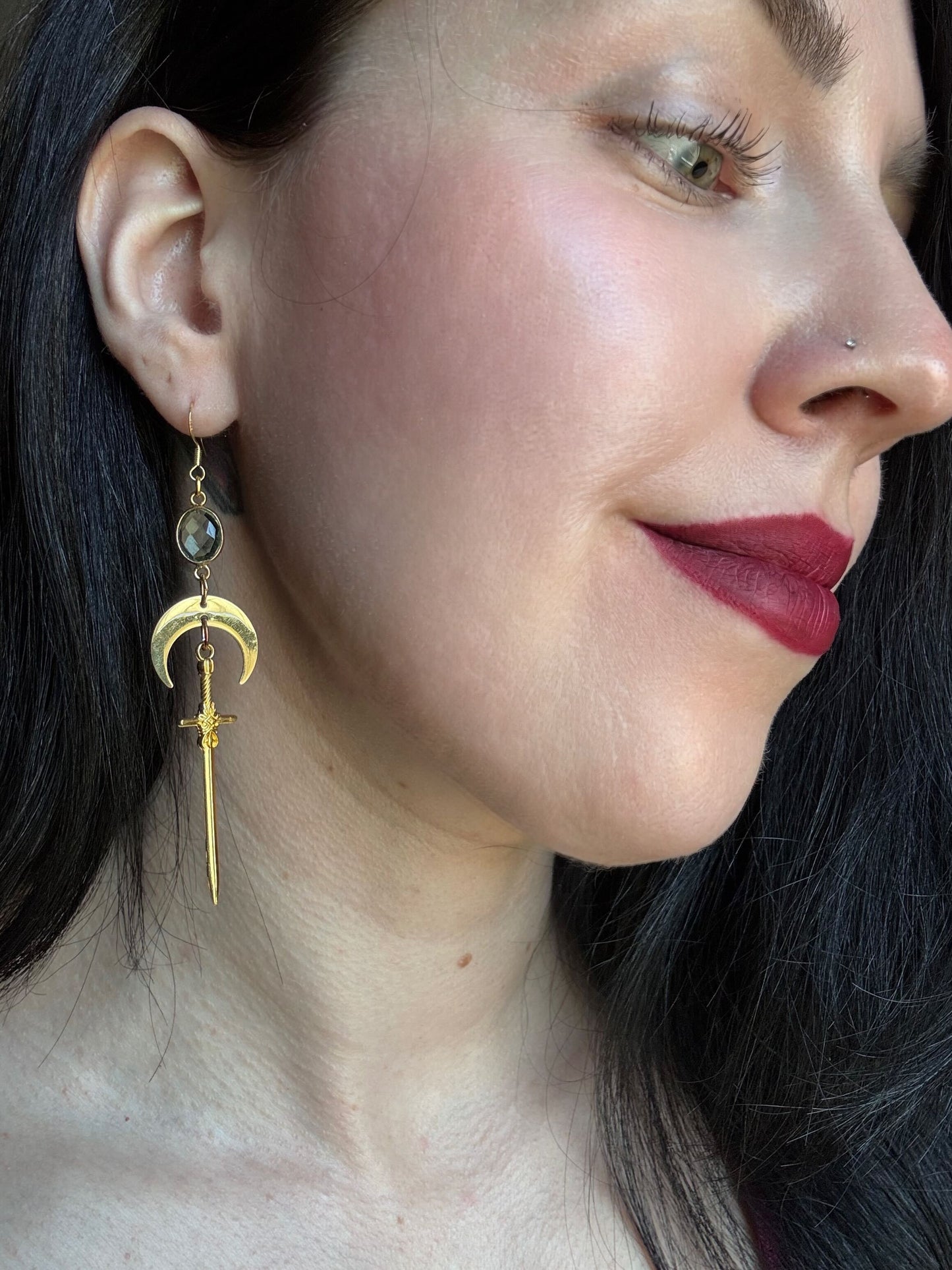 A woman wears a pair of green amethyst earrings with a gold moon and sword.