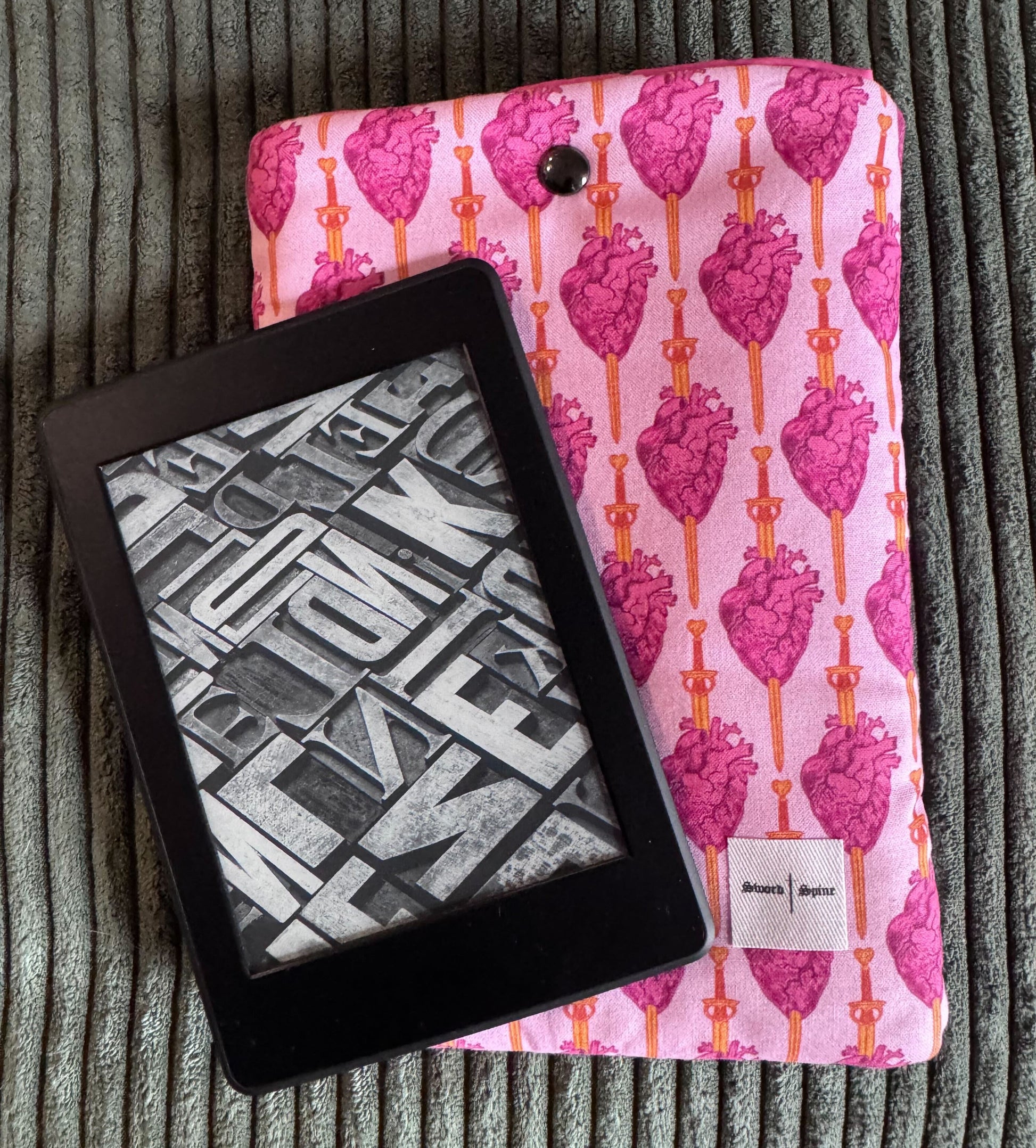 A kindle rests on a pink handmade e-reader sleeve with a sword piercing a heart pattern.