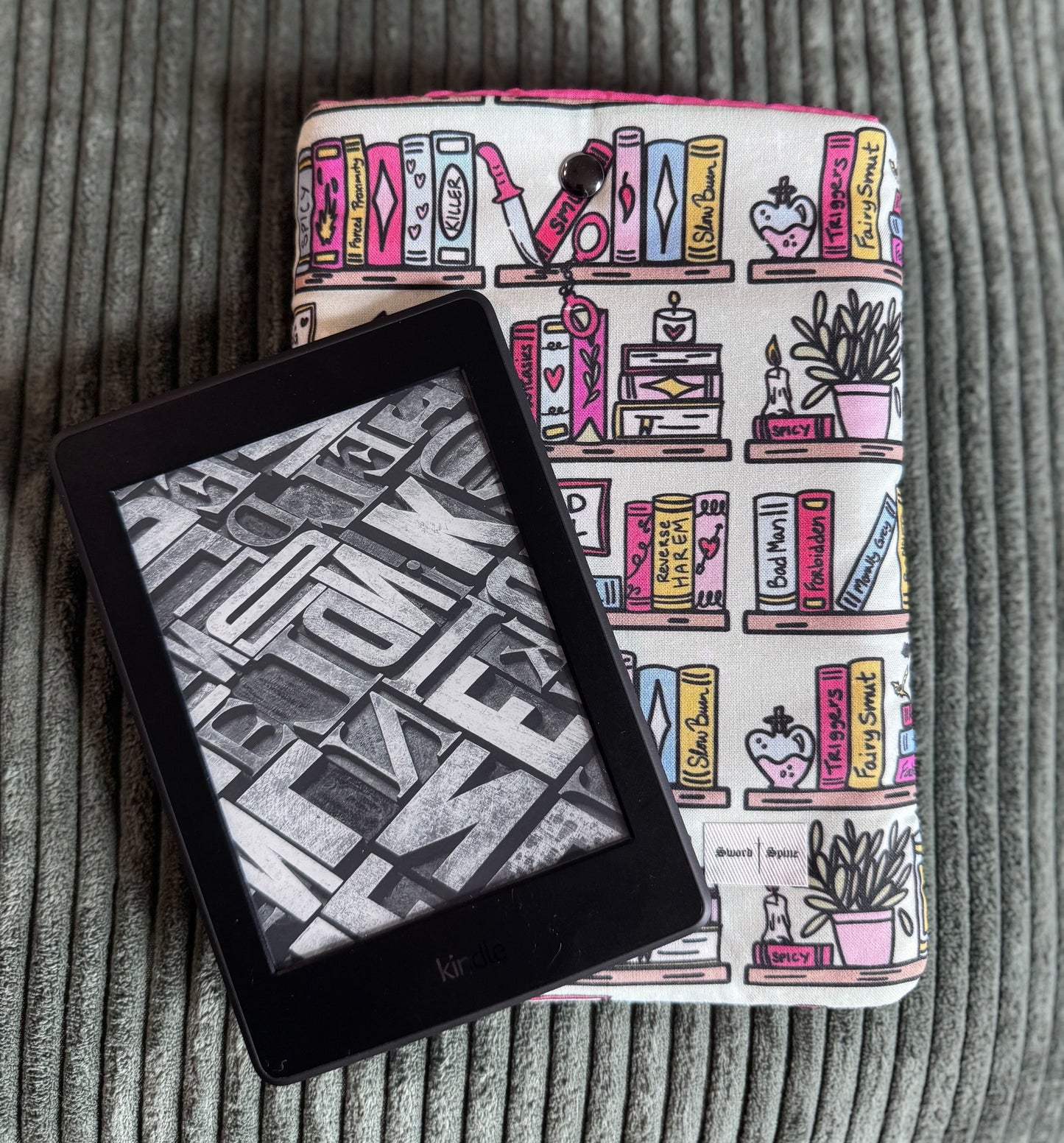 A kindle rests on top of a handmade e-reader sleeve with a pattern of romance books on shelves.