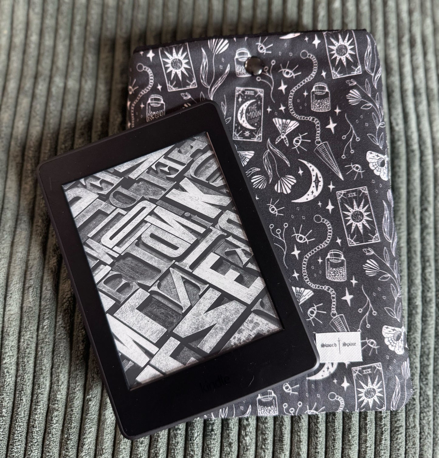 A kindle rests on top of a handmade e-reader sleeve with a black and white pattern of pendulums and tarot cards.