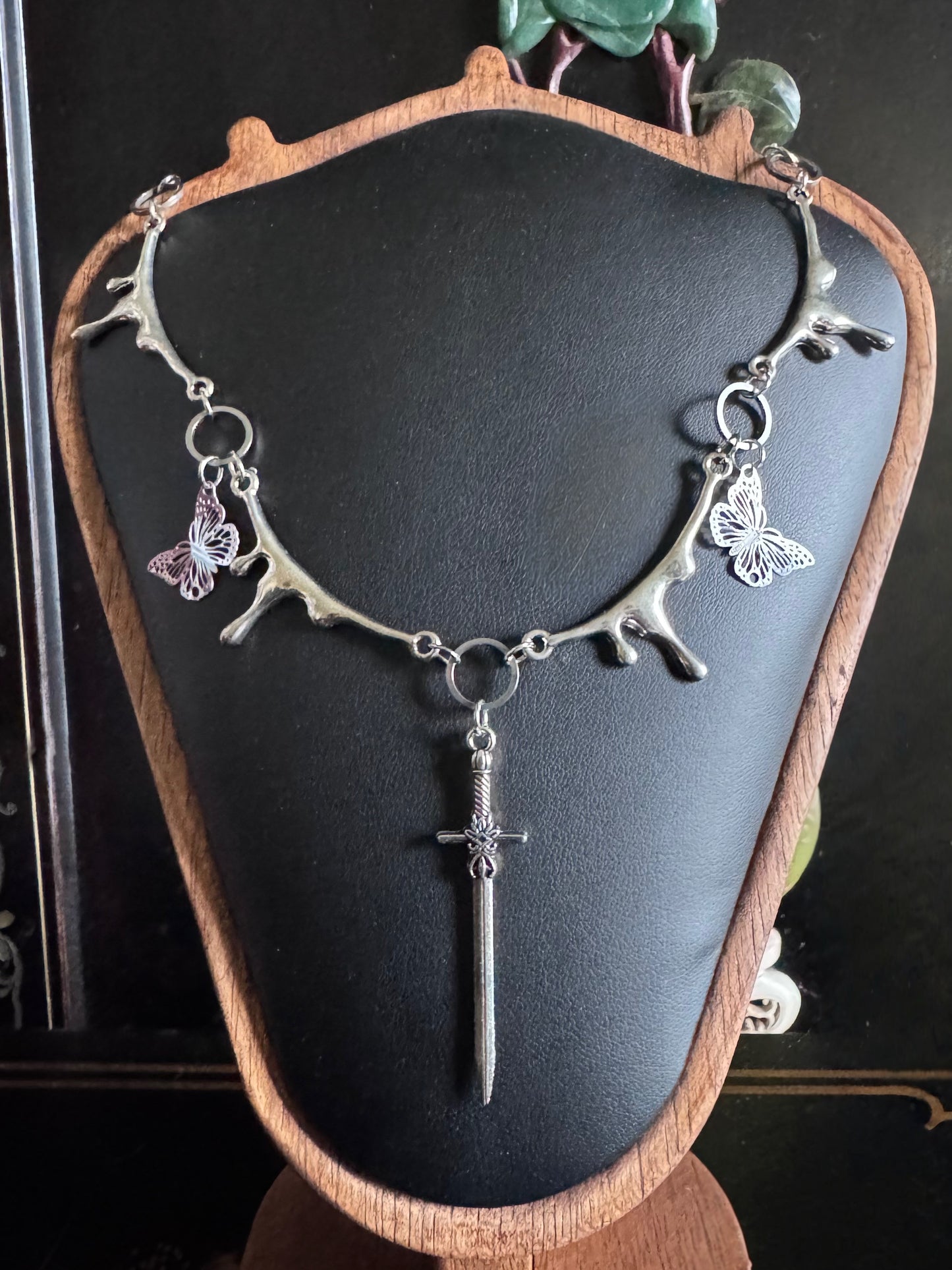 A dripping silver necklace with butterfly and sword pendant on a black mannequin bust.