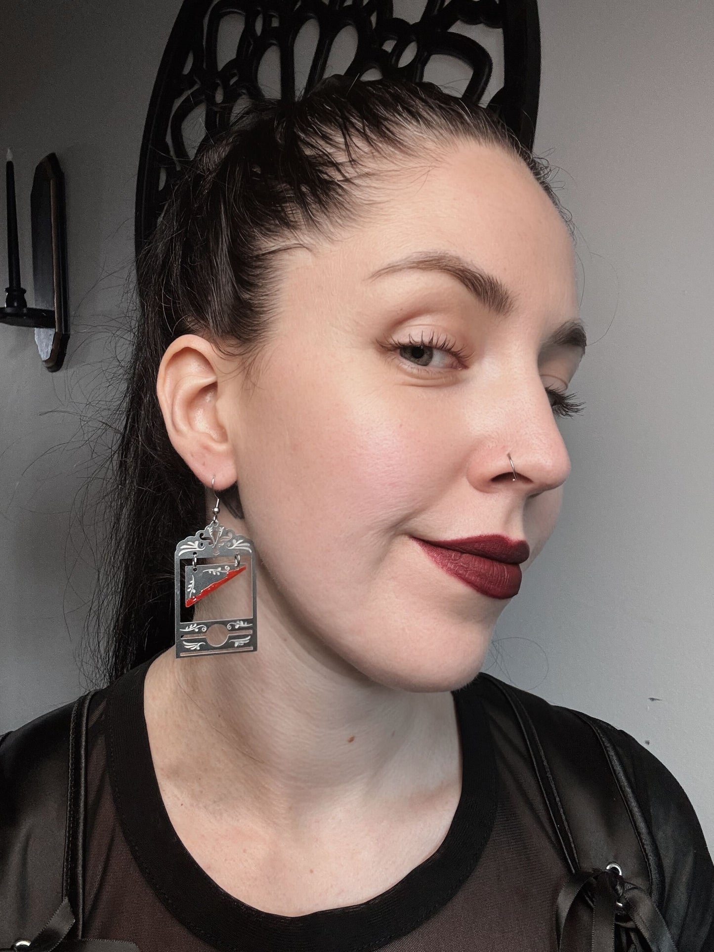 A woman with dark hair and red lips wears a pair of silver guillotine earrings that have red paint on the blade.