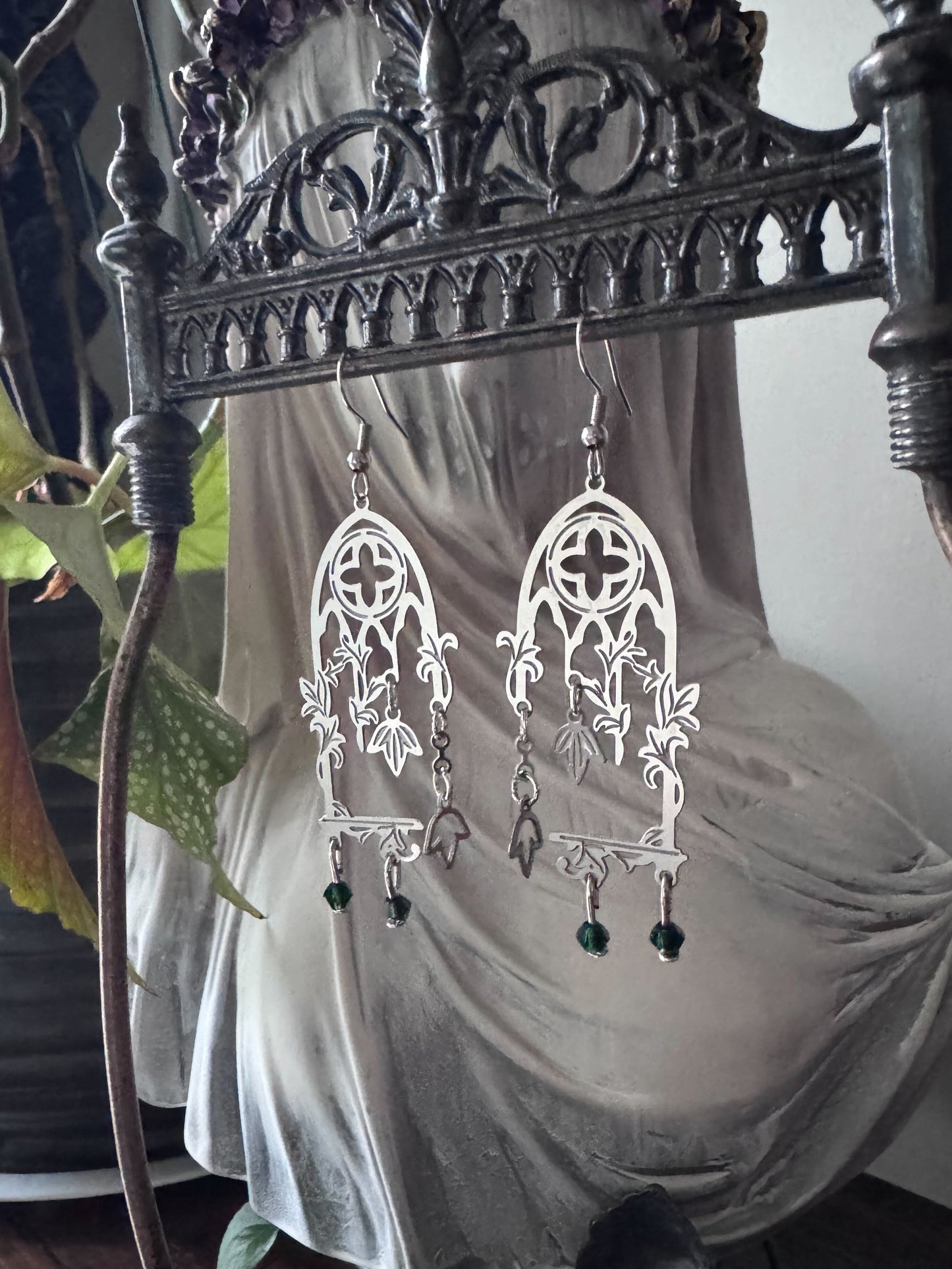 Decorative cathedral window earrings hanging on a gothic metal stand with a veiled bust and plant in the background.