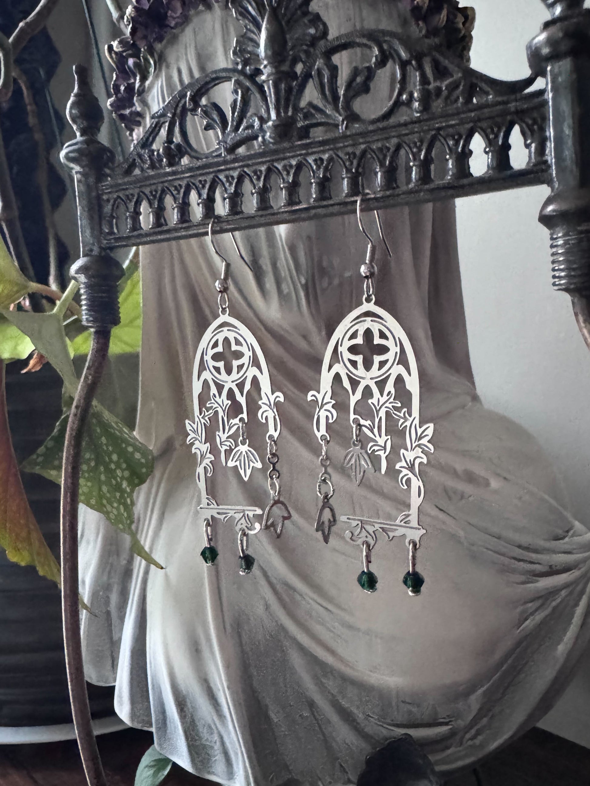 Decorative cathedral window earrings hanging on a gothic metal stand with a veiled bust and plant in the background.