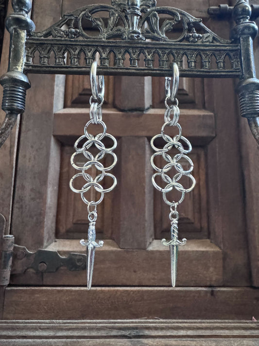 A pair of chainmail earrings with dagger pendants, hangs on an ornate display.
