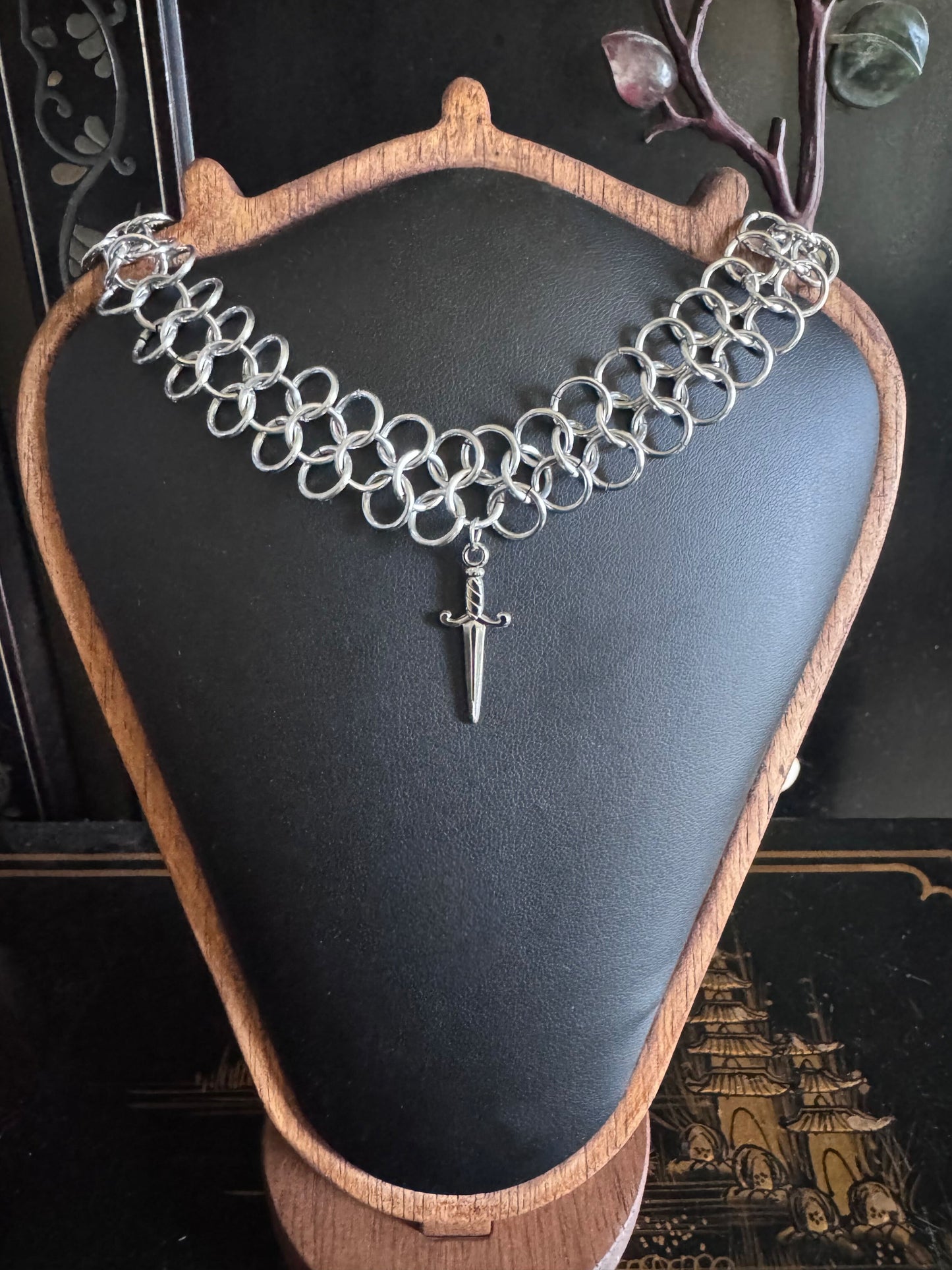 Silver chainmail necklace with a dagger pendant on a black display stand.