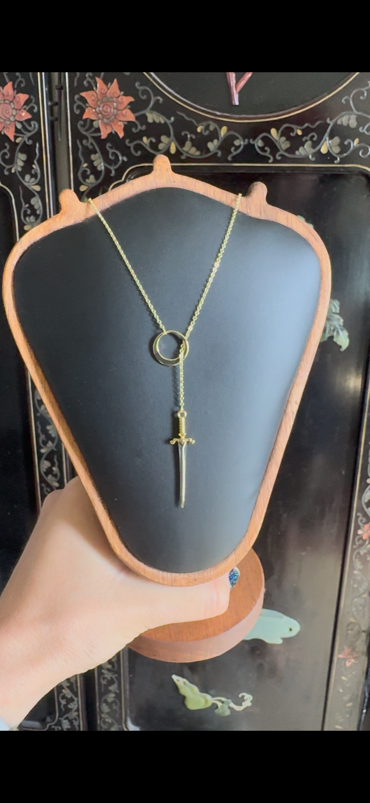 A lariat gold necklace with a sword through the loop, is on a black and wooden display.