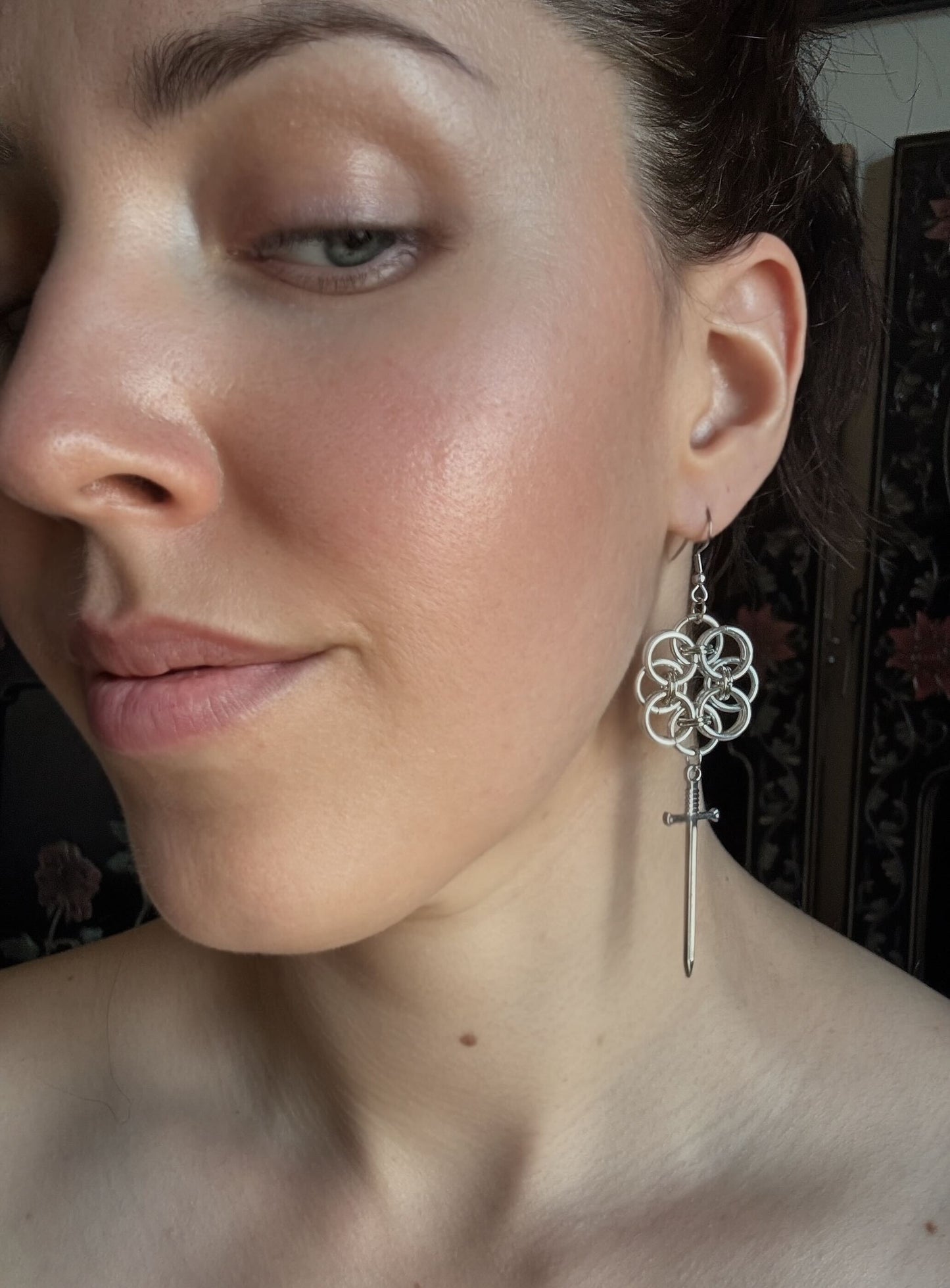 Close-up of a woman wearing a silver floral chainmail earring with a sword pendant.