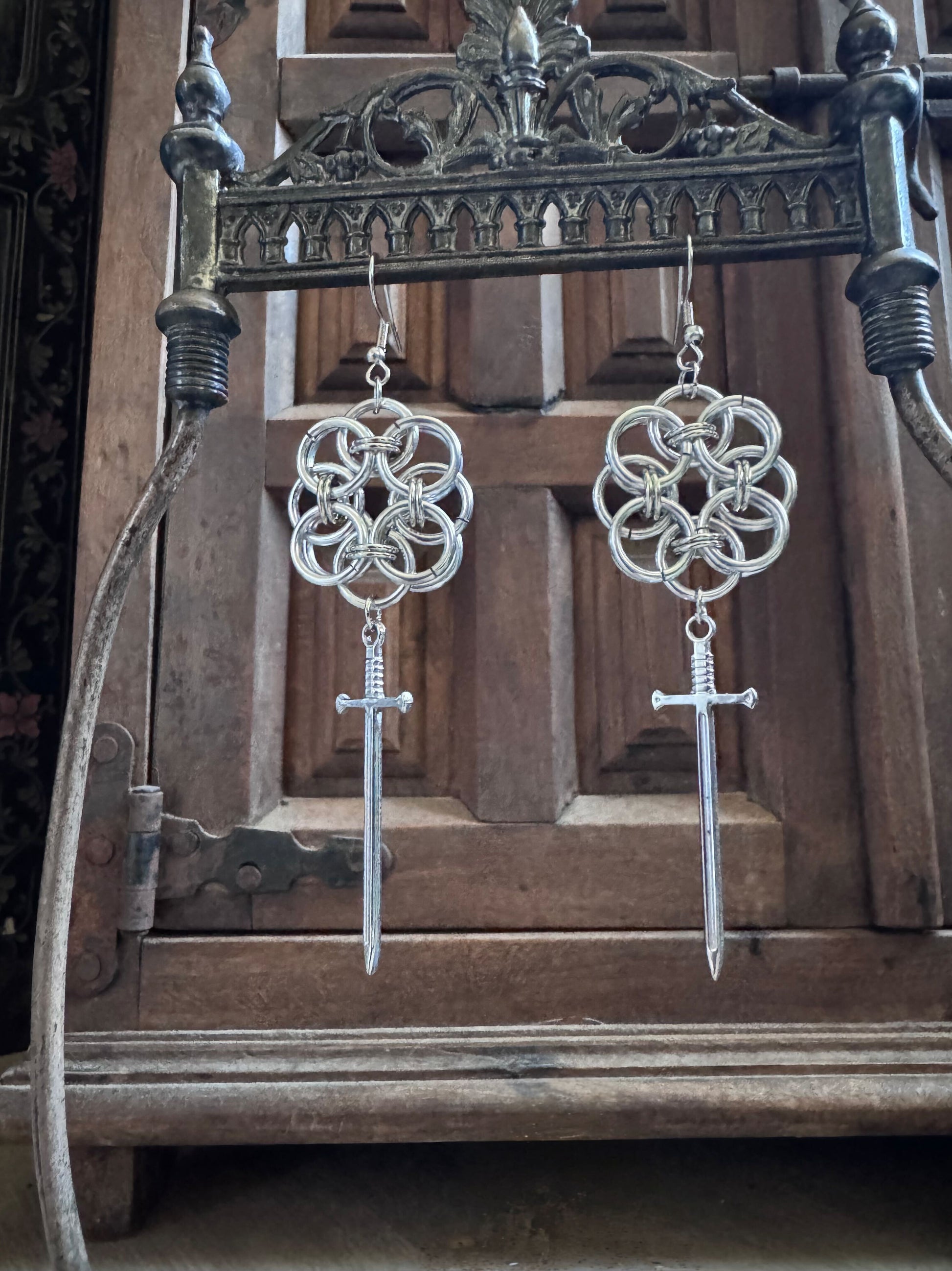 A pair of silver floral pattern chainmail earrings with sword pendants on an ornate metal display.