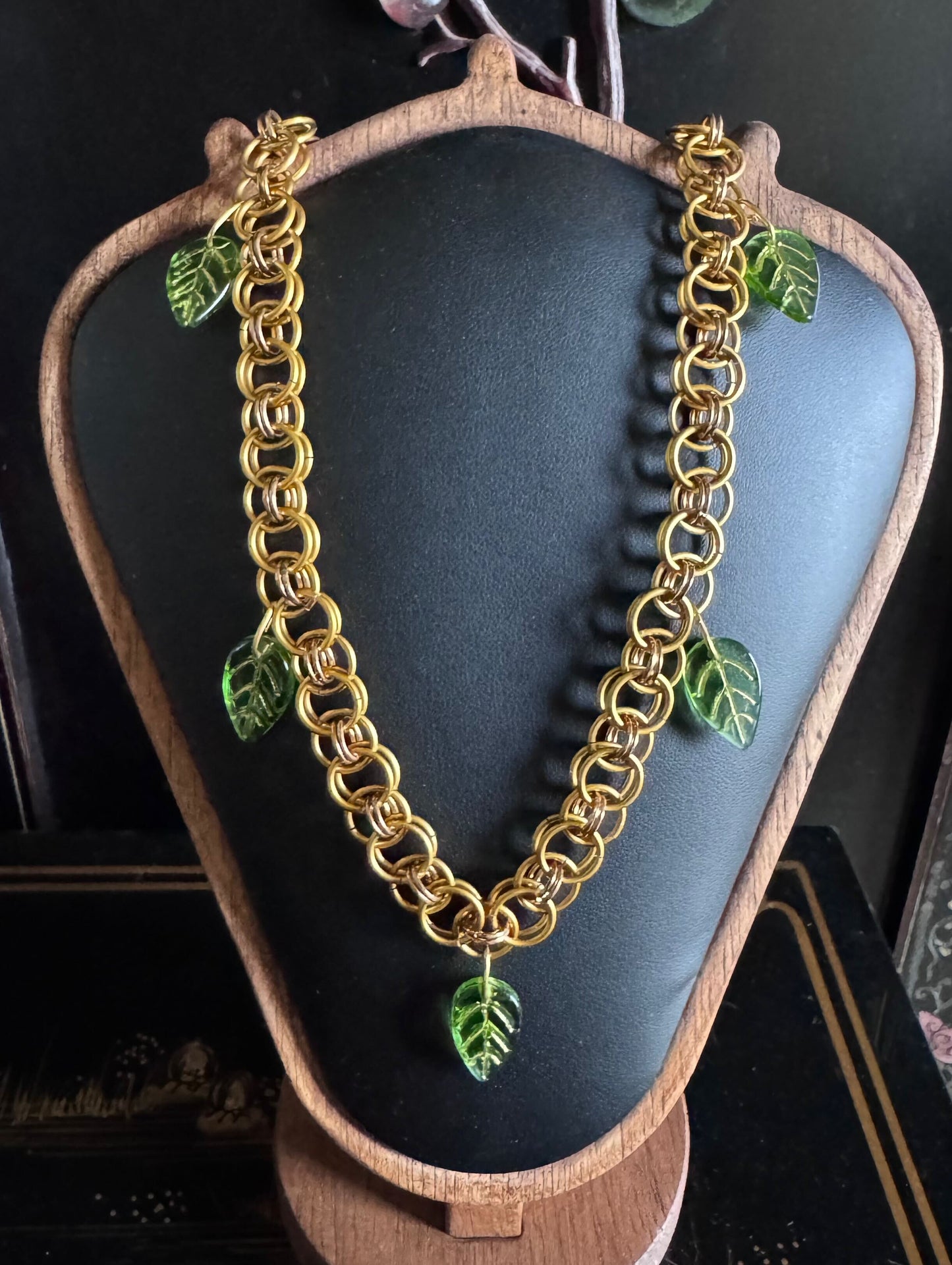 A beautiful gold chainmail necklace features a unique weave and green resin leaves attached throughout.