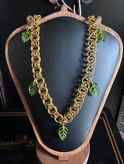 A beautiful gold chainmail necklace features a unique weave and green resin leaves attached throughout.