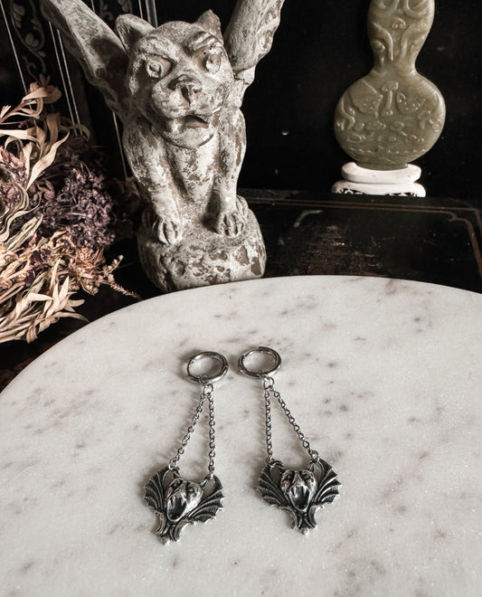 Pair of silver earrings with gargoyle designs on a marble surface, with decorative background elements.