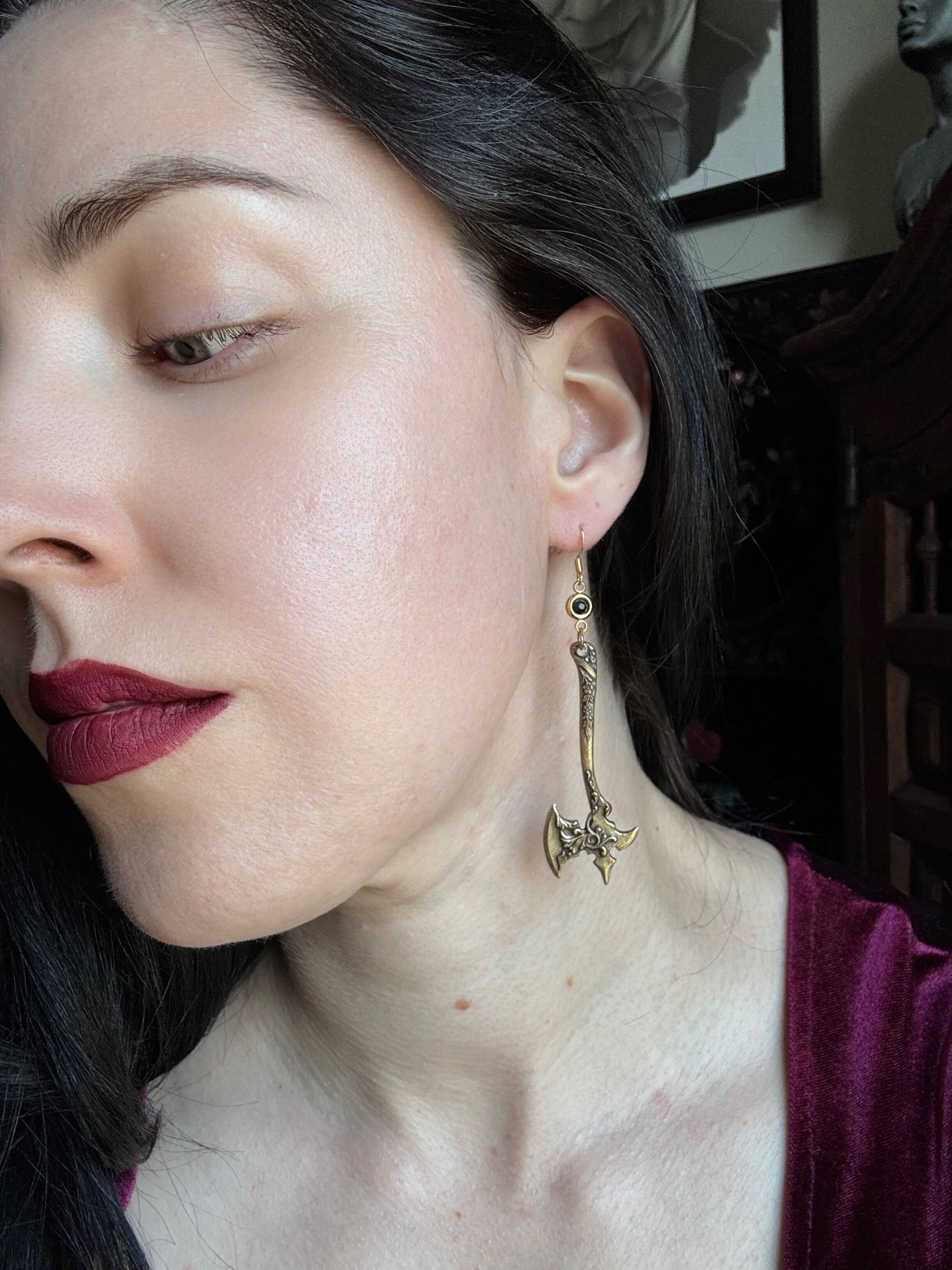 A woman wears a pair of golden brass medieval axe earrings with a black zirconia stone.