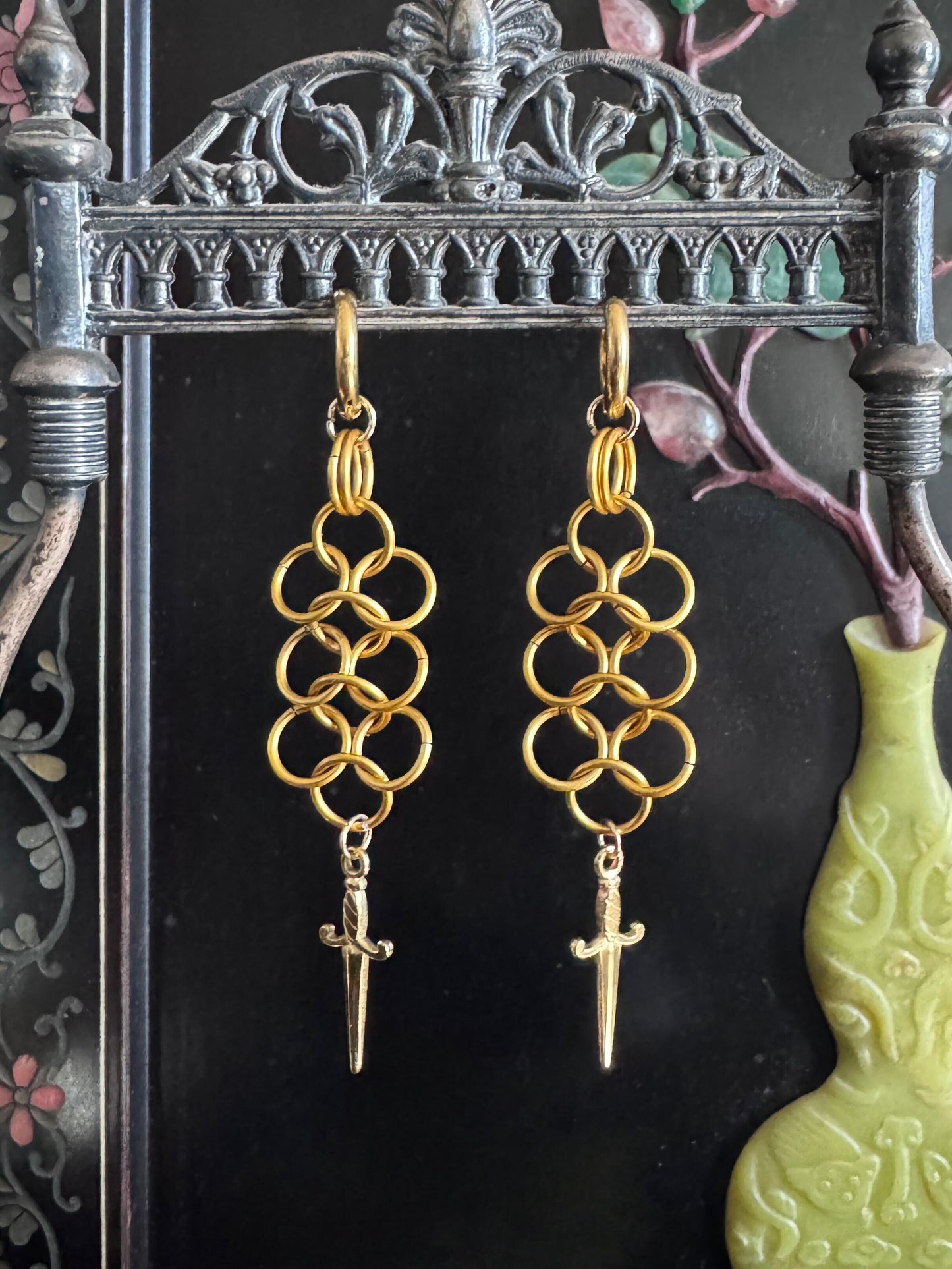 A pair of chainmail dangling earrings with daggers at the end of them, hang on a metal display.