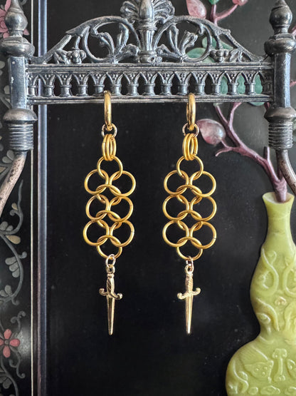 A pair of chainmail dangling earrings with daggers at the end of them, hang on a metal display.