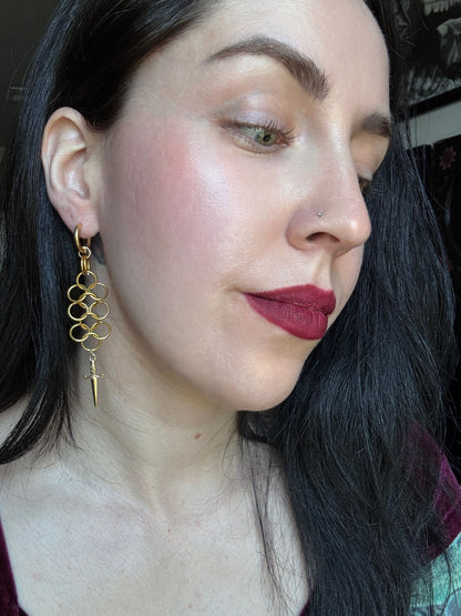 A woman wears a set of drop earrings featuring a chainmail weave and a dagger dangling from the bottom.