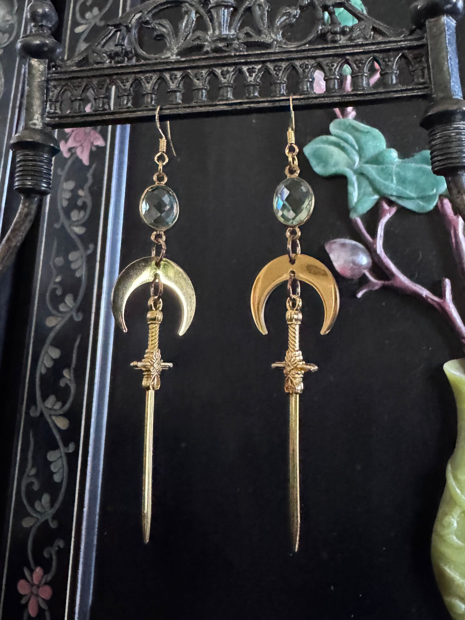 A pair of green amethyst earrings dangle with gold crescent moons and swords, creating a celestial fantasy effect.