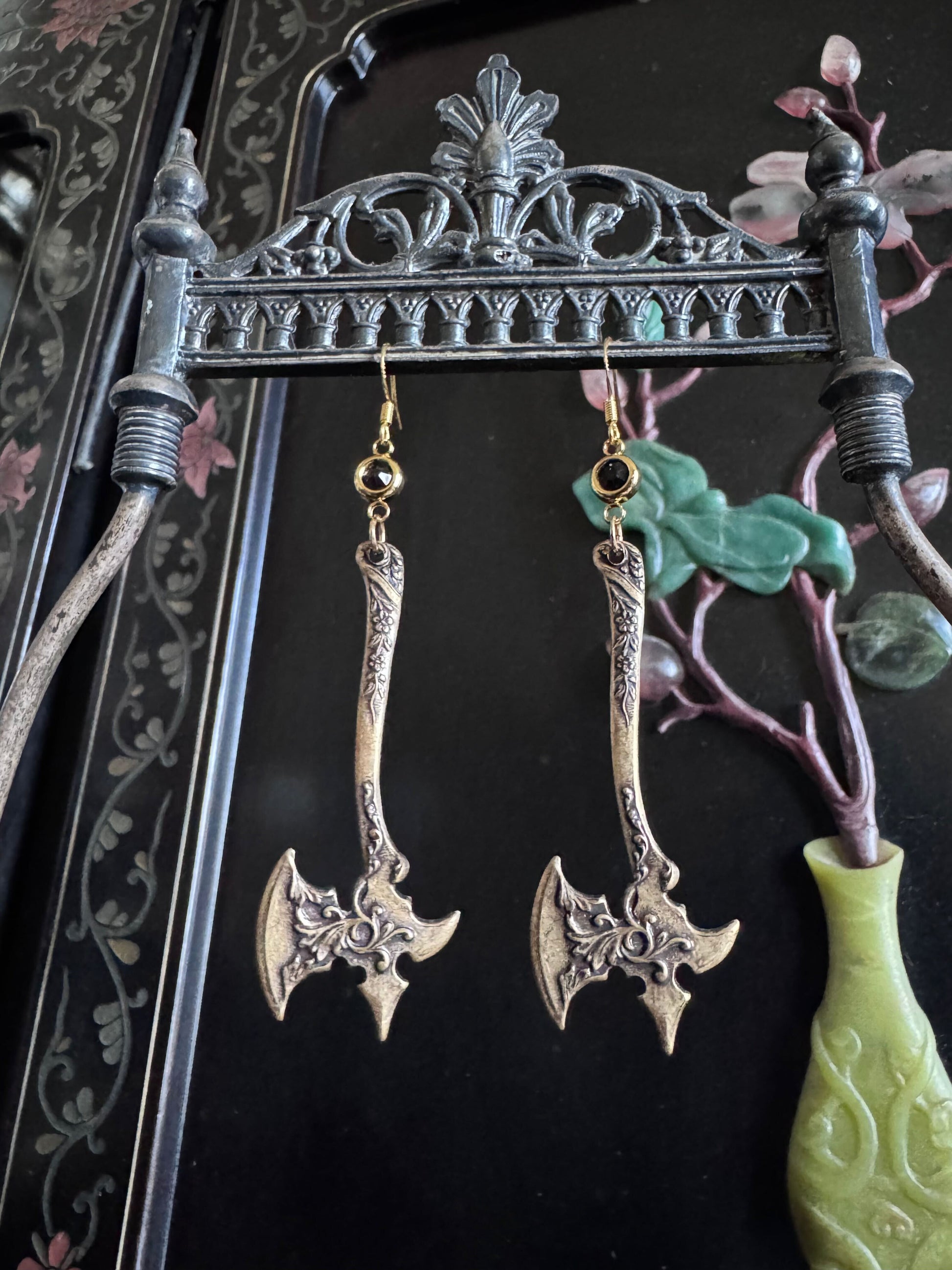 A pair of earrings featuring ornate detailed axes, dangle from black zirconia connectors on a metal display.