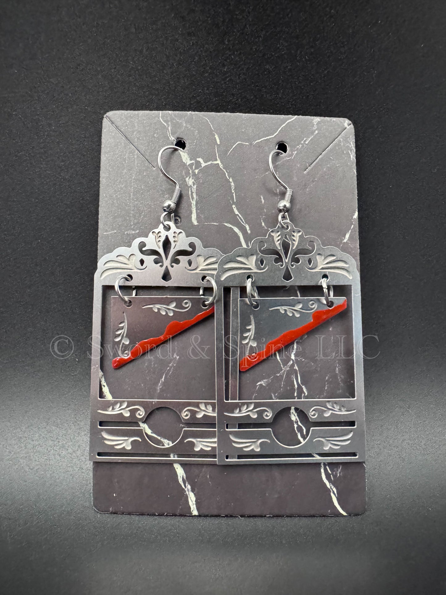 A pair of darkly elegant earrings with stainless steel guillotines featuring fake blood on the blade. 