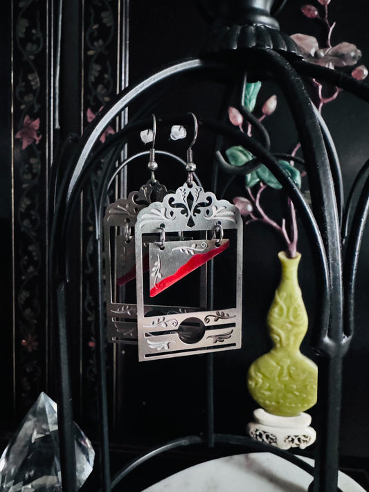 Silver guillotine earrings on a black metal stand with intricate designs.