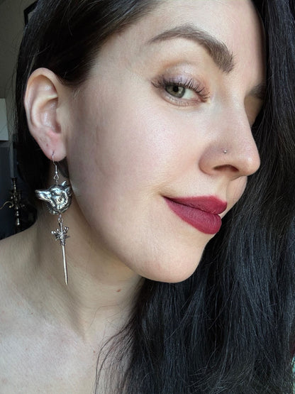 A woman wears a pair of silver wolf head earrings with a sword and crown dangling from them.