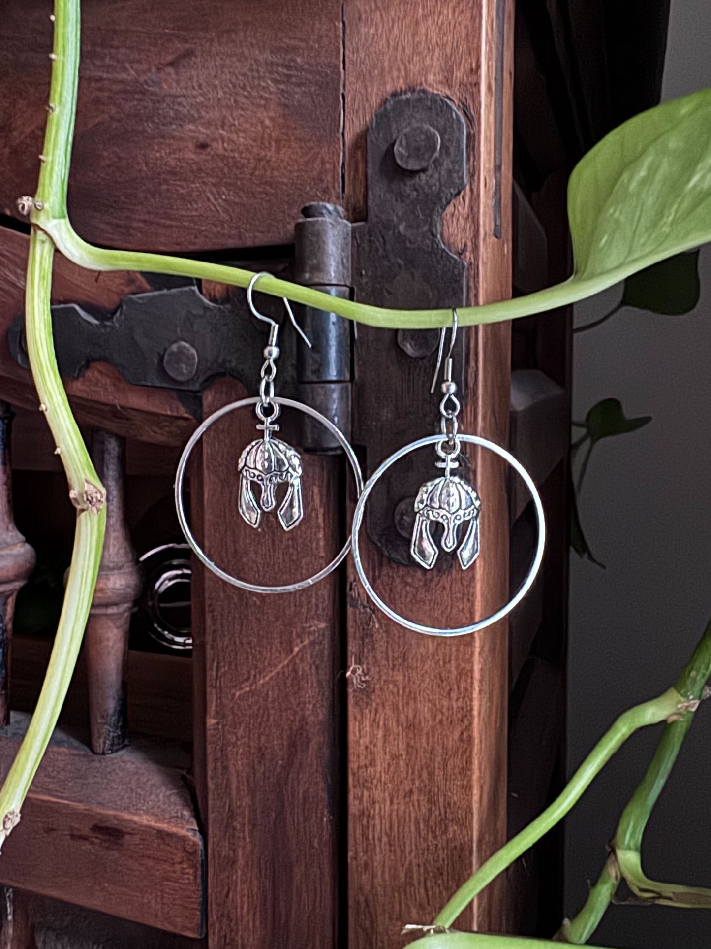 Silver hoop earrings with knight helmet charms hanging on a wooden surface surrounded by green leaves.