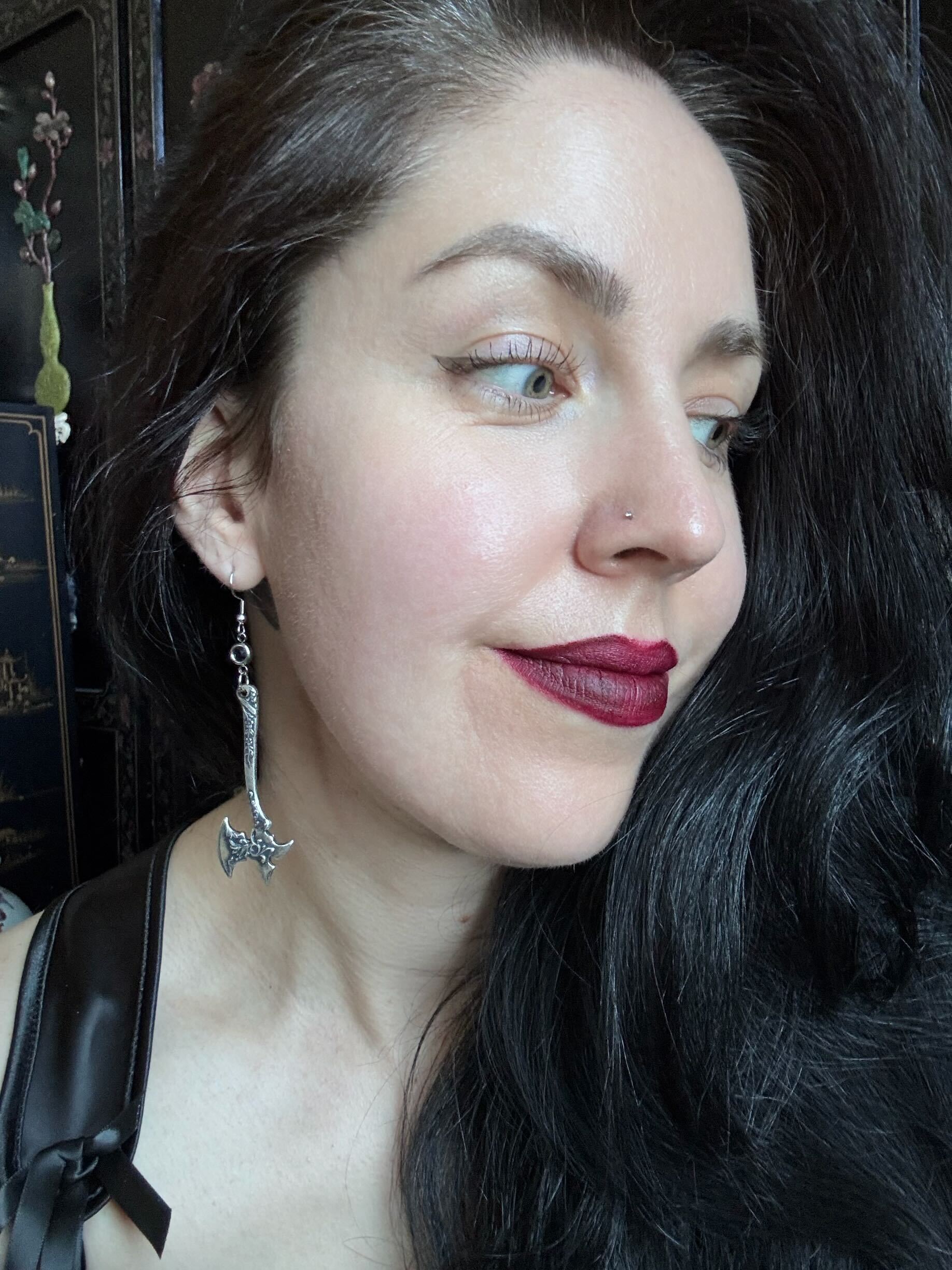 A woman with dark hair wears a pair of silver medieval axe earrings with ornate details.