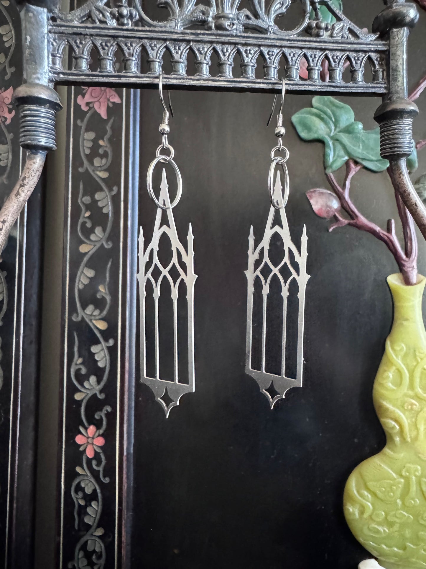 Gothic-style cathedral earrings hanging from a decorative frame with a vase and flowers in the background.