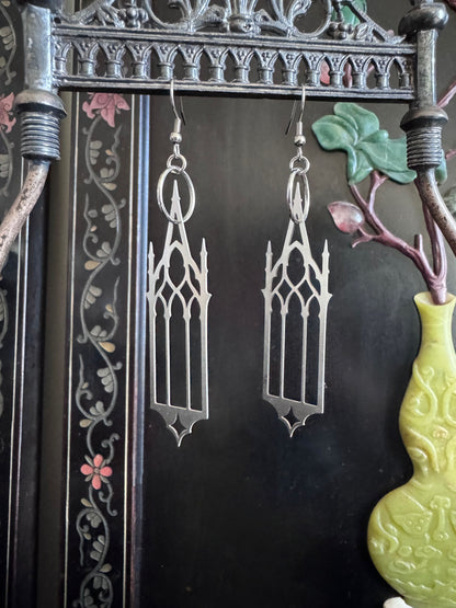 Gothic-style cathedral earrings hanging from a decorative frame with a vase and flowers in the background.