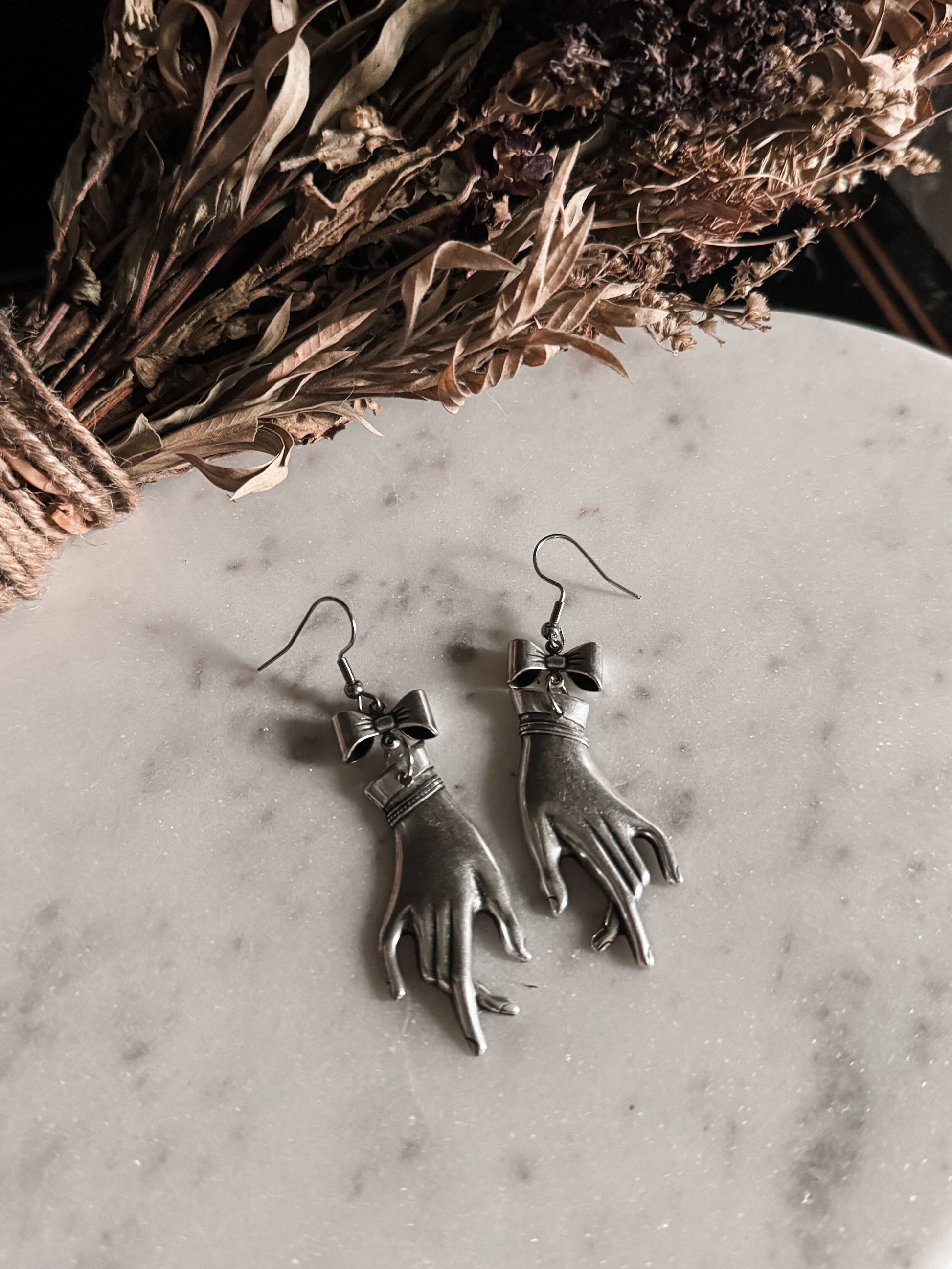 Silver Victorian mourning hand-shaped earrings on a marble surface with dried flowers in the background.