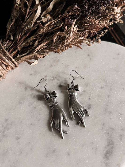 Silver Victorian mourning hand-shaped earrings on a marble surface with dried flowers in the background.