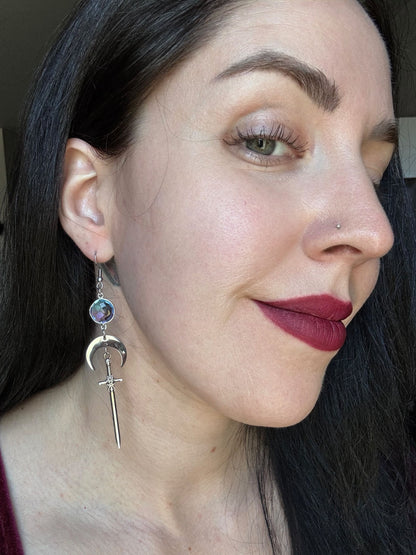 A woman wears purple blue ombre gemstone earrings with a crescent moon and sword.