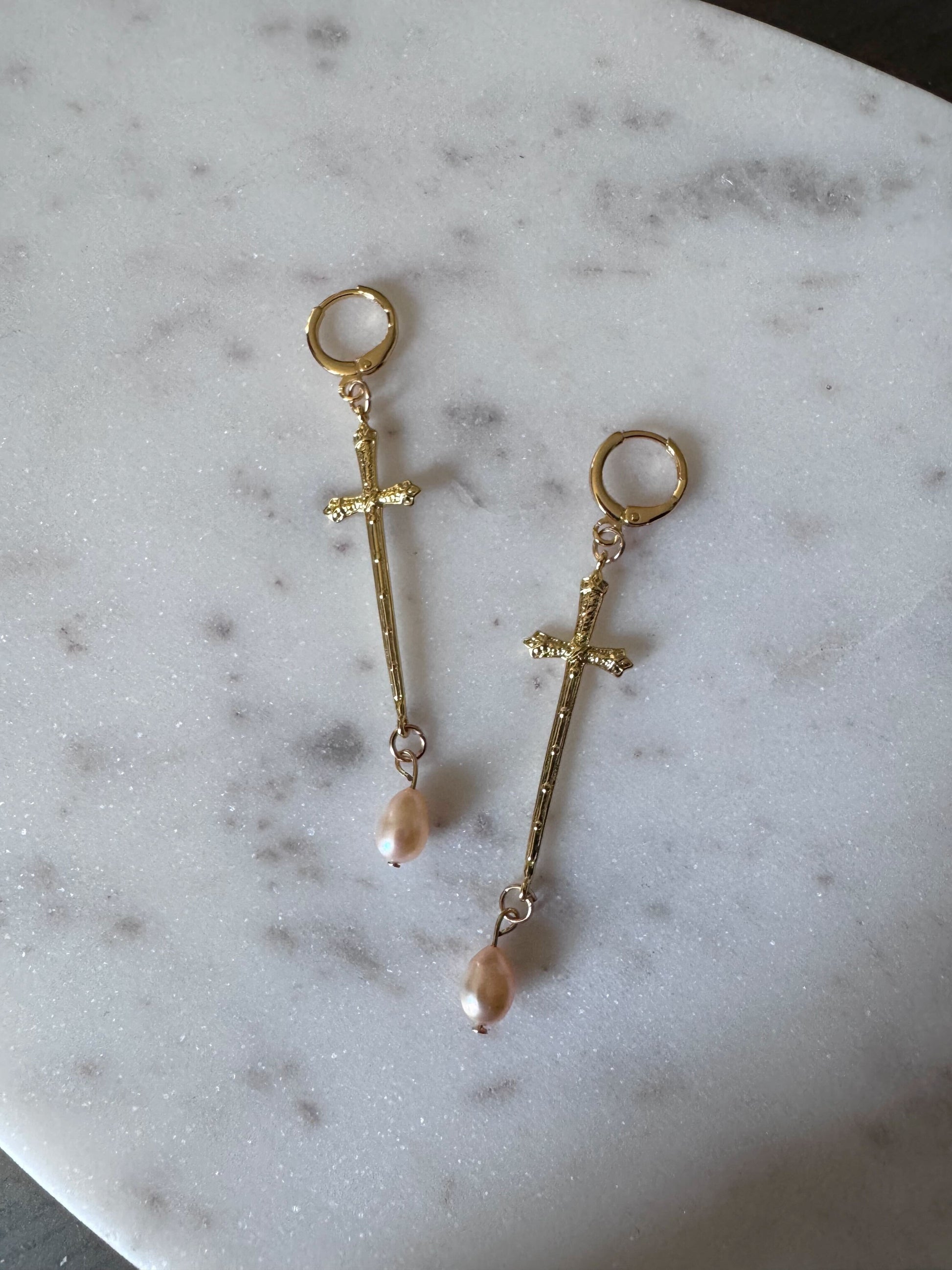 Golden cross like sword earrings with pink hued freshwater pearls on a marble surface.