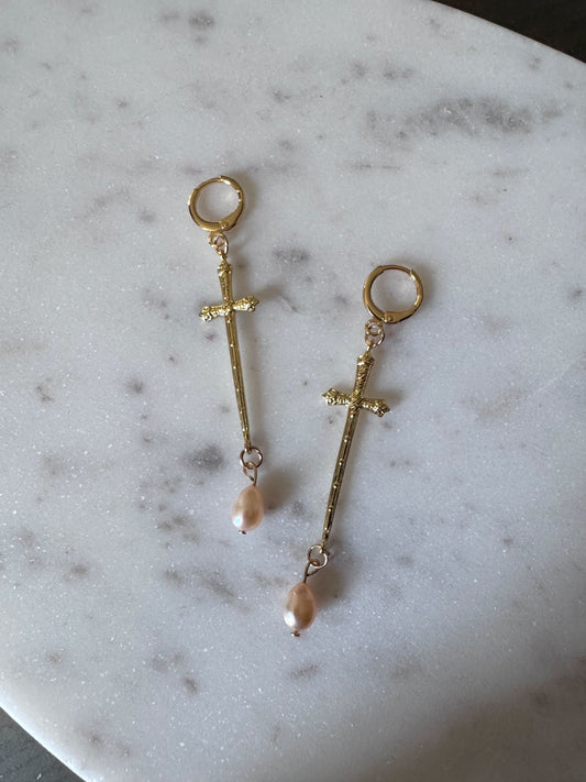 Golden cross like sword earrings with pink hued freshwater pearls on a marble surface.
