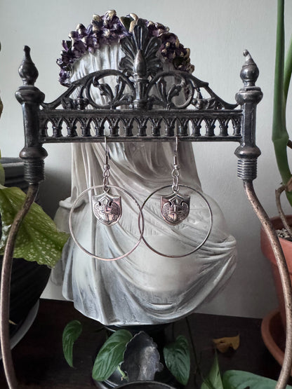 Silver hoop earrings with shield charms hanging from a gothic metal stand with plants in the background.