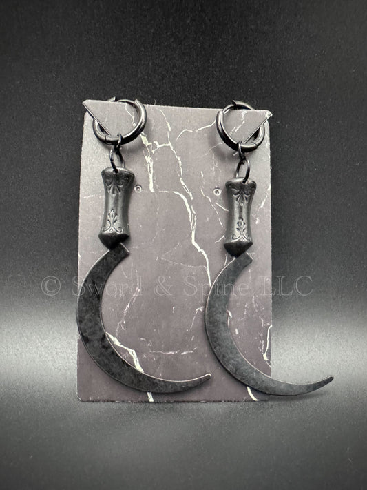 A set of black sickle/scythe earrings with black huggie clicker hoops displayed on a jewelry card.
