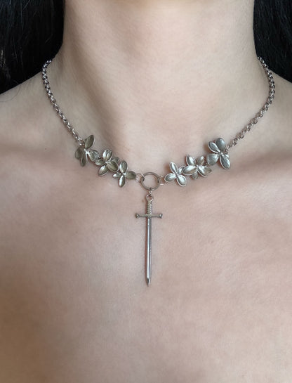 A silver necklace with cascading flowers on both sides, taper to a point with a stainless steel sword pendant.