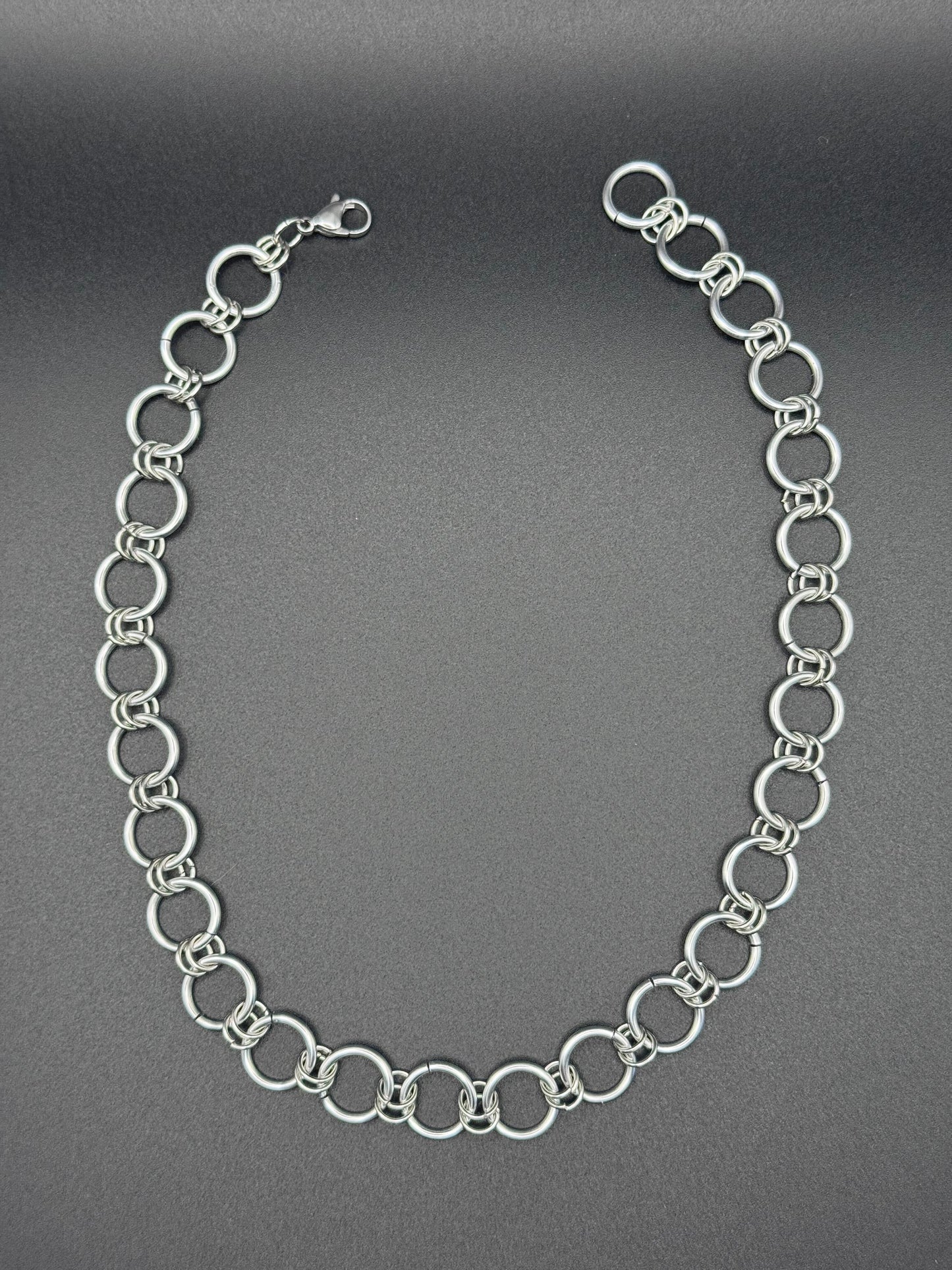 A simple and elegant silver chainmail necklace rests against a dark background.