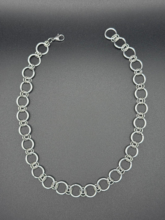 A simple and elegant silver chainmail necklace rests against a dark background.