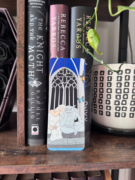 A hand drawn laminated bookmark rests on a bookshelf and features Sybil and Bartholomew from the fantasy romance best seller The Knight and The Moth.