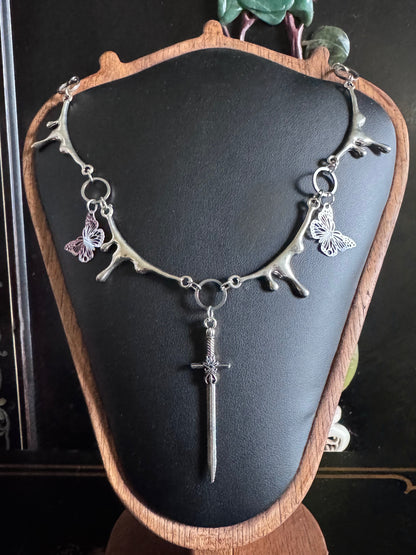 A dripping silver necklace with butterfly and sword pendant on a black mannequin bust.