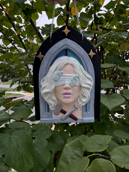 A hand painted bas relief of a woman with her eyes covered by a white cloth, a hammer and chisel at the base of her neck, set against a gothic arch with stars in the background.