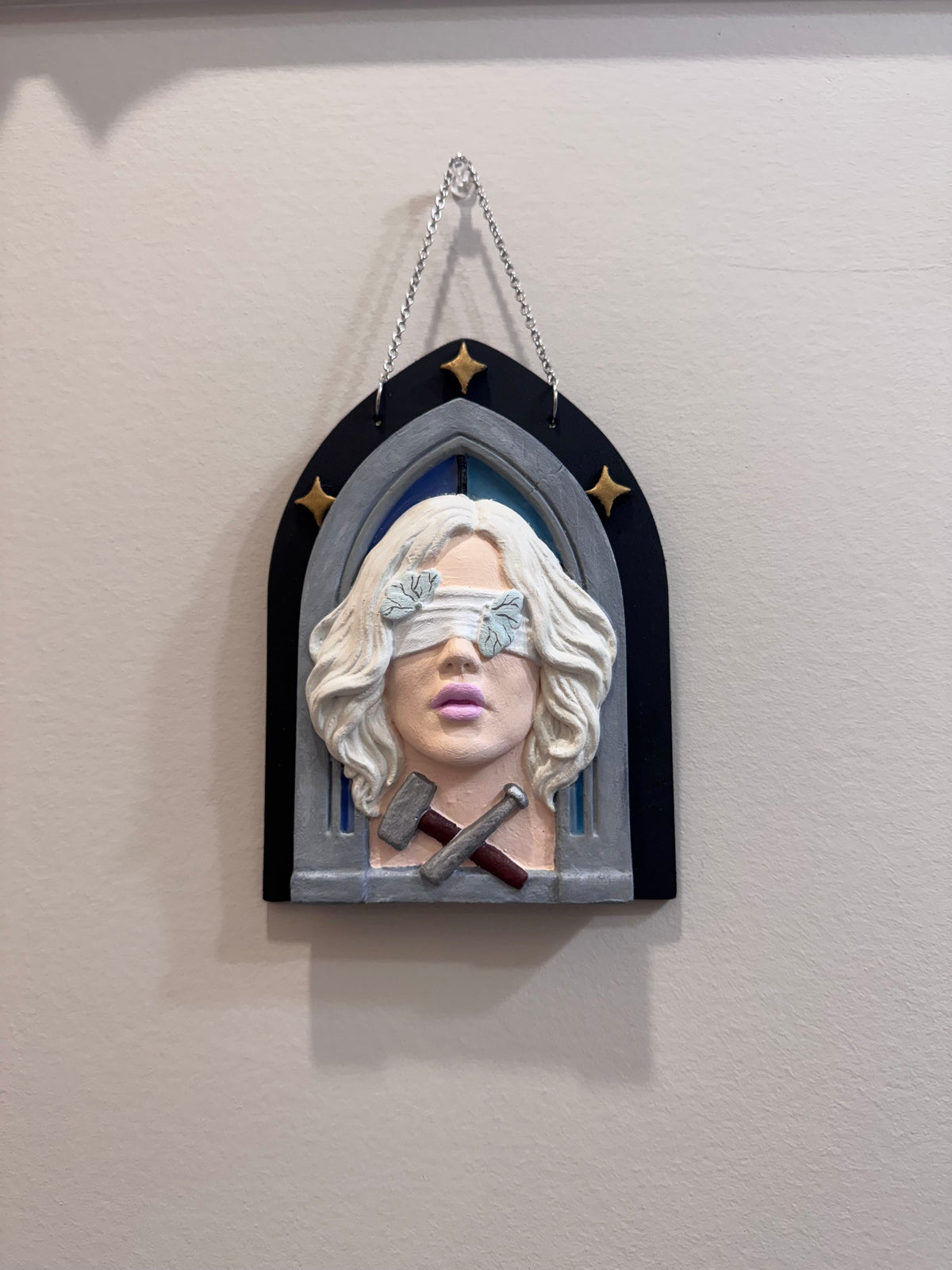 A hand painted bas relief of a woman with a hammer and chisel at her collarbone, set against a gothic arch with stars in the background.