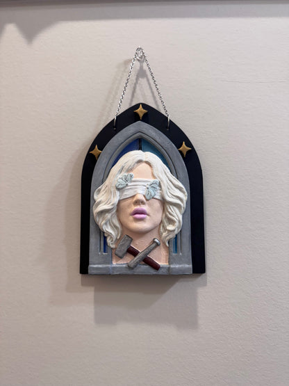A hand painted bas relief of a woman with a hammer and chisel at her collarbone, set against a gothic arch with stars in the background.
